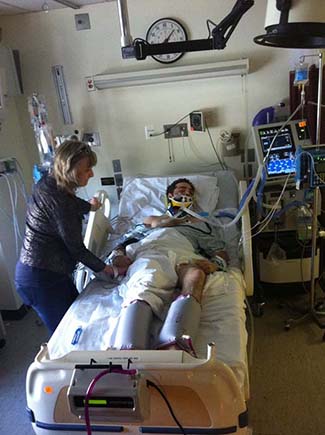 Cavin's mother stands at his hospital bedside, holding his hand. Cavin is laying down, unconscious, hooked up to various tubes and machinery to keep him in stable condition.