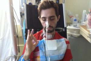 Cavin sits in a wheelchair next to his hospital bed, eyes closed, giving the "OK" sign with his hand. He has a tracheostomy and a breathing apparatus.