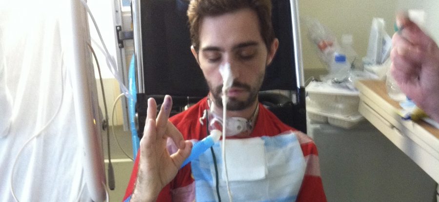 Cavin sits in a wheelchair next to his hospital bed, eyes closed, giving the "OK" sign with his hand. He has a tracheostomy and a breathing apparatus.