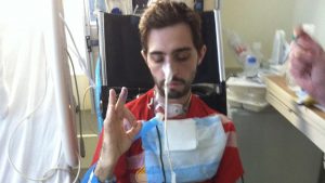 Cavin sits in a wheelchair next to his hospital bed, eyes closed, giving the "OK" sign with his hand. He has a tracheostomy and a breathing apparatus.